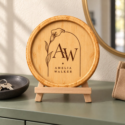 A round wooden plaque or coaster with engraved initials 'AW' and the name 'Amelia Walker' beneath it, displayed on a small wooden stand on a tabletop in a styled interior setting.