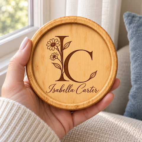 A hand holding a round wooden coaster engraved with a floral monogram and the name 'Isabella Carter' in a warm indoor setting.